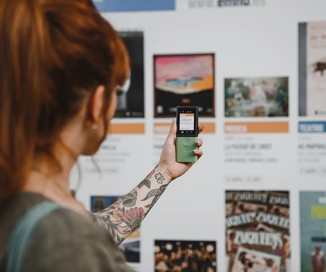 Asmuo su raudonais plaukais ir gėlių tatuiruote ant rankos laiko žalią skaitmeninį įrenginį, nukreipdamas jį į vitriną su įvairiais plakatais ir informacija apie įvykį.