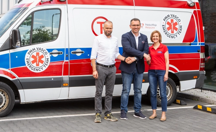 Trys žmonės stovi ir šypsosi priešais baltą, raudoną ir mėlyną greitosios pagalbos automobilį, stovintį asfaltuotoje gatvėje šalia pastato. Du vyrai, vienas su kostiumu, kitas su baltais marškiniais, spaudžia vienas kitam ranką, prie jų prisijungia moteris su raudonais marškiniais.