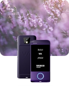 Produkto pristatymo nuotrauka, kurioje vaizduojamas violetinis „Vasco Translator“ įrenginys iš dviejų kampų prieš tamsų, tekstūruoto audinio foną. Kairėje pusėje esantis įrenginys parodytas iš nugaros pusės, akcentuojant fotoaparato objektyvą su blykste ir reljefinį „VA“ logotipą. Dešinėje pusėje esantis įrenginys rodomas iš priekio su įjungtu ekranu, kuriame matomi sveikinimai „Hello!“, „VA“, „¡Hola!“ ir „VASCO Translator“ logotipas. Apvalus mygtukas įrenginio priekinės dalies apačioje apibrėžtas šviečiančia mėlyna šviesa. Kompozicija išdėstyta ant svajingo, neryškaus fono iš violetinės viržės gėlių, apšviestų švelnia saulės šviesa.