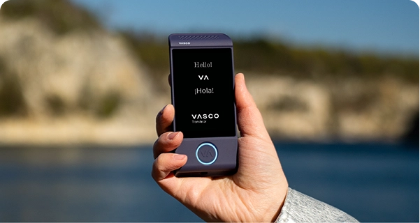 Artimas rankos, laikanti violetinį elektroninį vertimo įrenginį „Vasco“, kadras prieš ryškų lauko foną. Ekrane matomi sveikinimai „Hello!“, „¡Hola!“ ir „VASCO Translator“ logotipas. Fone – švelniai sulietintas vaizdas saulėto pakrantės peizažo su mėlynu vandeniu ir šviesios spalvos uolėtomis uolomis po giedru dangumi. Asmuo, laikantis įrenginį, dėvi šviesiai pilkos arba sidabrinės spalvos tekstūruotą striukės rankovę. 