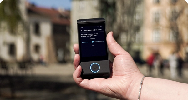 Artimas žmogaus rankos, kurioje laikomas juodas elektroninis vertėjas Vasco, kadras prieš neryškų lauko foną – miesto aikštę su istoriniais pastatais. Prietaiso ekrane rodomi „Vertimo skaitymo greičio“ nustatymai: slankiklis nustatytas į padėtį „Normalus“, o šalia yra užrašas: „Pakeiskite vertimo skaitymo greitį, perkelkdami slankiklį.“ Asmuo, laikantis prietaisą, nešioja sidabrinę apyrankę. Ryškus dienos šviesos spindulys išryškina ekrano sąsają, todėl nustatymus lengva matyti. 