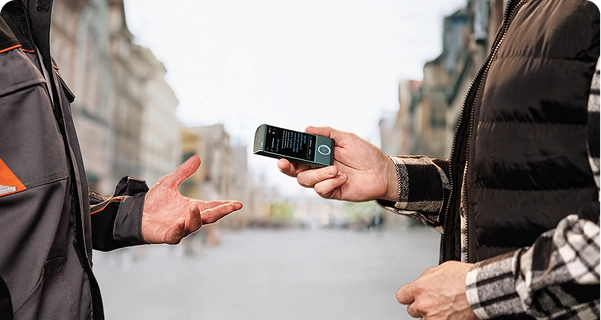Žmogus, vilkintis juodą dygsniuotą liemenę ant languotos marškinių, miesto gatvėje horizontaliai laiko žalsvai mėlyną Vasco elektroninį vertėją, nukreiptą į kitą asmenį. Antrasis asmuo, vilkintis pilką ir oranžinę darbo striukę, ištiesęs ranką tarsi kalbėtųsi. Prietaiso ekrane matomas vertimo langas su tekstu. Fone – platus, neryškus vaizdas į europietiško stiliaus gatvę su aukštais pastatais po ryškiai apsiniaukusiu dangumi. 
