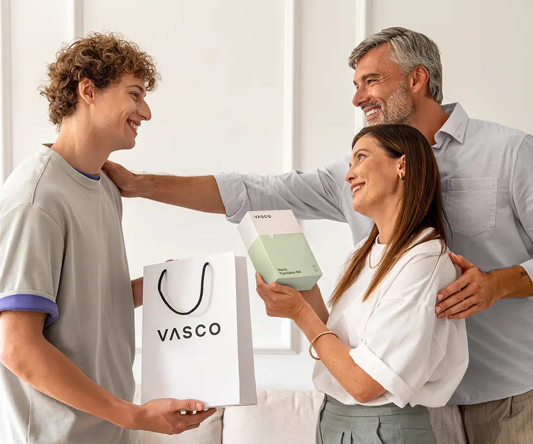 Širdį šildanti scena, kurioje trys žmonės stovi kartu ir šypsosi. Kairėje – garbanotais plaukais jaunuolis, rankoje laikantis baltą „VASCO“ pirkinių maišelį. Centre – moteris, laikanti baltą ir šviesiai žalią dėžutę su užrašu Vasco Translator M4 ir laimingai žiūrinti į jaunuolį. Už jos stovi vyras su žilais plaukais, šiltai šypsosi, viena ranka padėjęs ant moters peties, o kita – ant jauno vyro peties. Jie stovi šviesioje patalpoje su neutralios spalvos sienomis, o fone matosi sofa. 