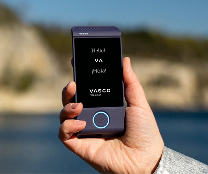 Artimas rankos, laikanti violetinį elektroninį vertimo įrenginį Vasco Translator M4, kadras prieš ryškų lauko foną. Ekrane matomi sveikinimai „Hello!“, „¡Hola!“ ir „VASCO Translator“ logotipas.